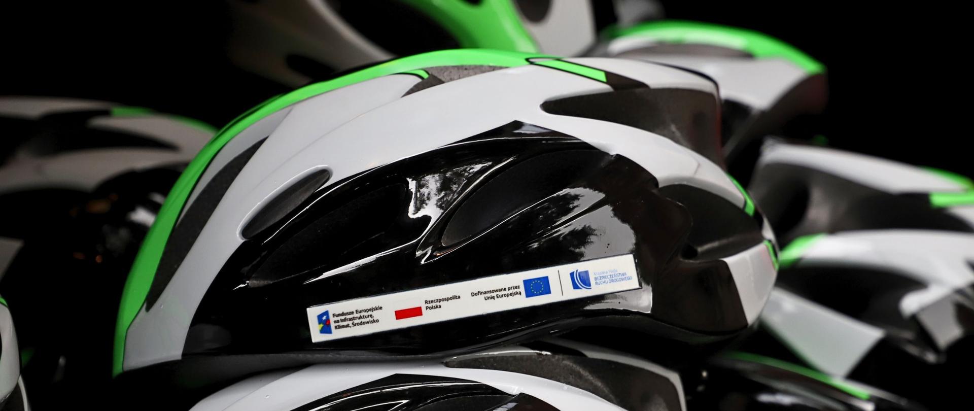 Kask zielono-biało-czarny z naklejonymi logotypami: Fundusz Europejski na Infrastrukturę, Klimat i Środowisko, Rzeczypospolita Polska, dofinansowane przez Unię Europejską, Krajowa Rada Bezpieczeństwa Ruchu Drogowego. W tle większa ilość kasków.