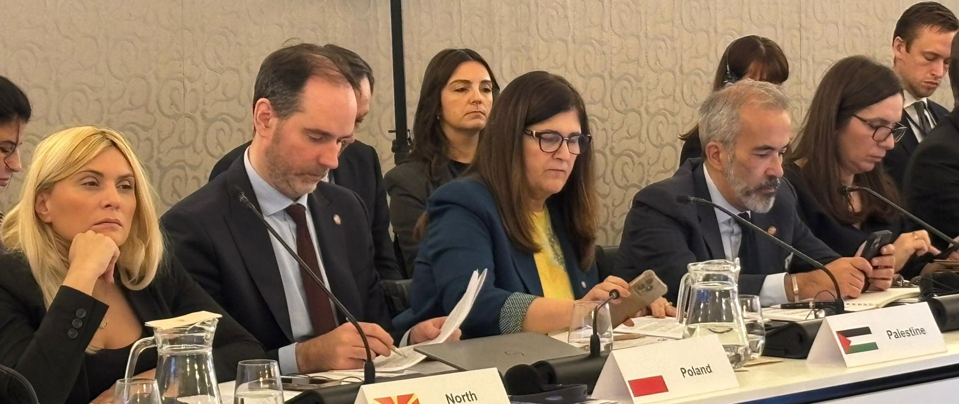 Na I planie przy długim stole konferencyjnym siedzi 5 osób w formalnych ubraniach. Mężczyzna od lewej strony patrzy w kartkę i coś notuje.