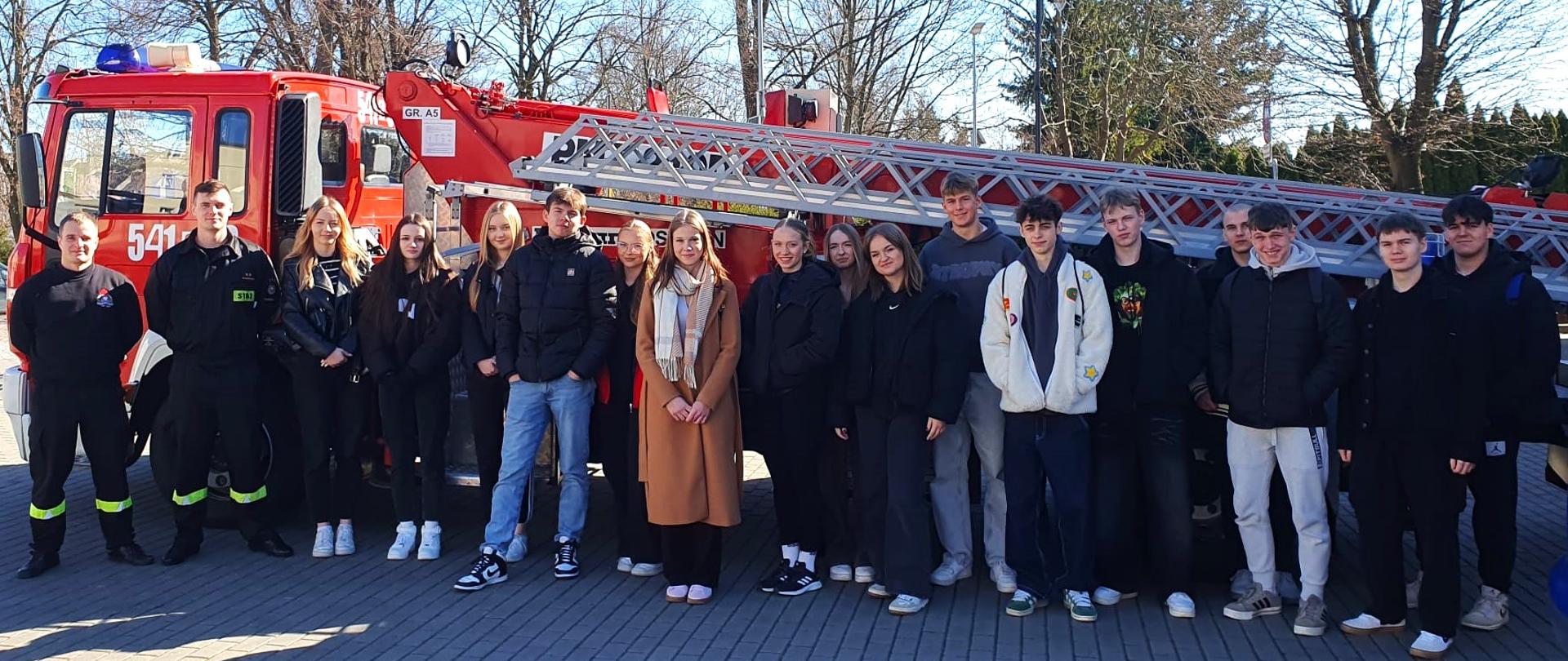 Grupa osób stoi na placu. Z lewej strony stoi dwóch strażaków. Strażacy mają na sobie czarne umundurowanie. Obok strażaków stoi grupa młodzieży. Za grupą ludzi stoi wóz strażacki. Wóz strażacki jest koloru czerwonego. Na wozie strażackim są małe elementy koloru białego. Na wozie strażackim widoczna jest drabina. Drabina jest koloru szarego. W tle widać drzewa i bezchmurne niebieskie niebo. 