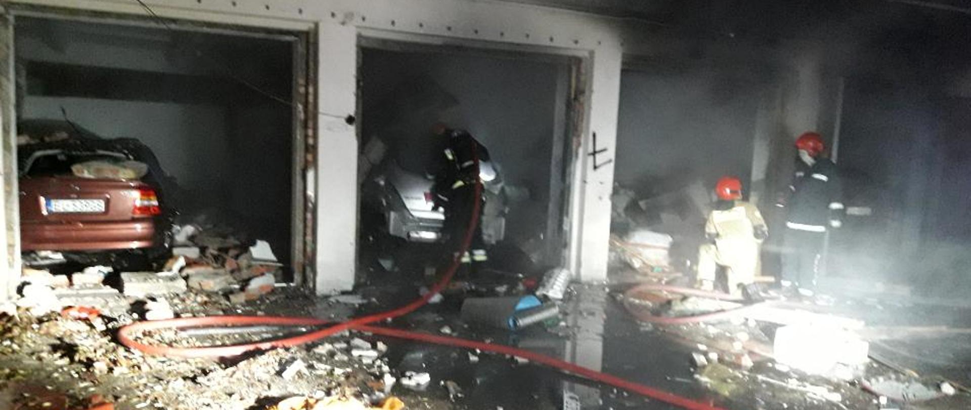 Zdjęcie przedstawia kompleks garaży po wybuchu, uszkodzone bramy garażowe, oraz strażaków prowadzących akcję gaśniczą.