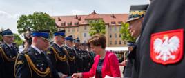Na zdjęciu widać odznaczonych i wyróżnionych funkcjonariuszy PSP w mundurach wyjściowych. Zdjęcie wykonane w ciągu dnia. W tle widać krzewy i drzewa z zieloną koroną. Za drzewami, w centralnym punkcie budynek.