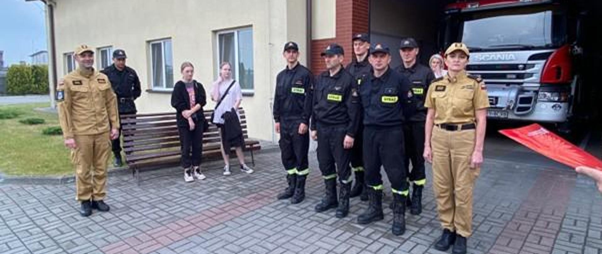 Na pierwszym planie widzimy funkcjonariuszy, którzy ubrani są w umundurowanie służbowe oraz gości, którzy uczestniczyli w uroczystej zmianie służby, gdyż wylicytowali voucher na spędzenie jednego dnia ze zmianą służbową w Jednostce Ratowniczo-Gaśniczej w Rypinie. Na drugim planie natomiast widzimy garaże komendy i wóz gaśniczy. 