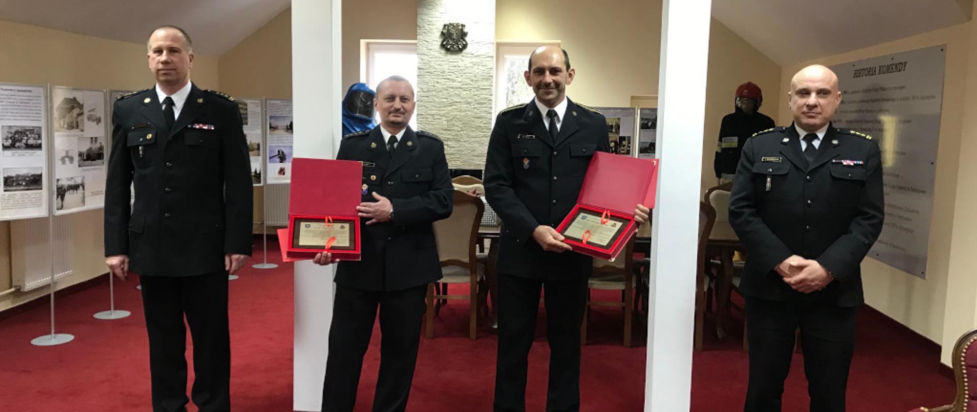 Zdjęcie przedstawia czterech strażaków w umundurowaniu galowym. Dwaj strażacy znajdujący się w środku trzymają w rękach listy gratulacyjne. Po lewej stronie znajduje się Komendant Powiatowy Państwowej Straży Pożarnej w Jędrzejowie, natomiast po lewej stronie- Zastępca Komendanta. Wydarzenie ma miejsce w sali tradycji, w której znajdują się różne tablice z wydarzeniami historycznymi tutejszej komendy.