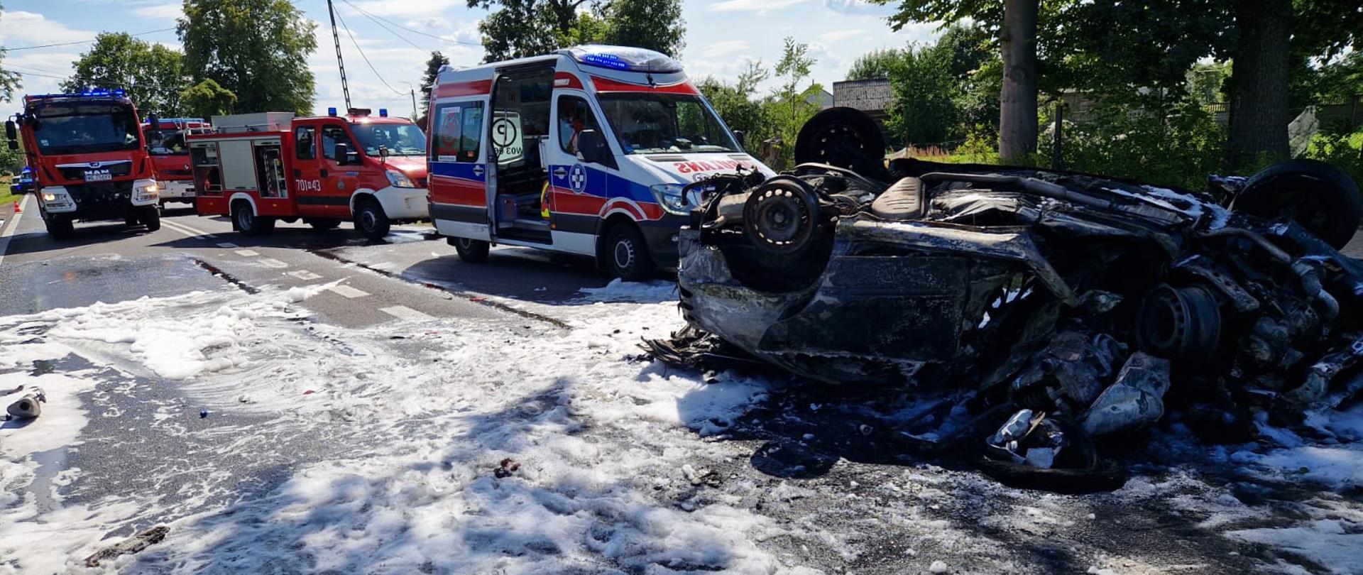 Motocykl wbity w samochód osoby, pojazdy uległy spaleniu. Samochód osobowy leży na dachu. W tle widać pojazdy ratownicze.