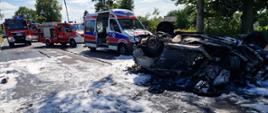 Motocykl wbity w samochód osoby, pojazdy uległy spaleniu. Samochód osobowy leży na dachu. W tle widać pojazdy ratownicze.