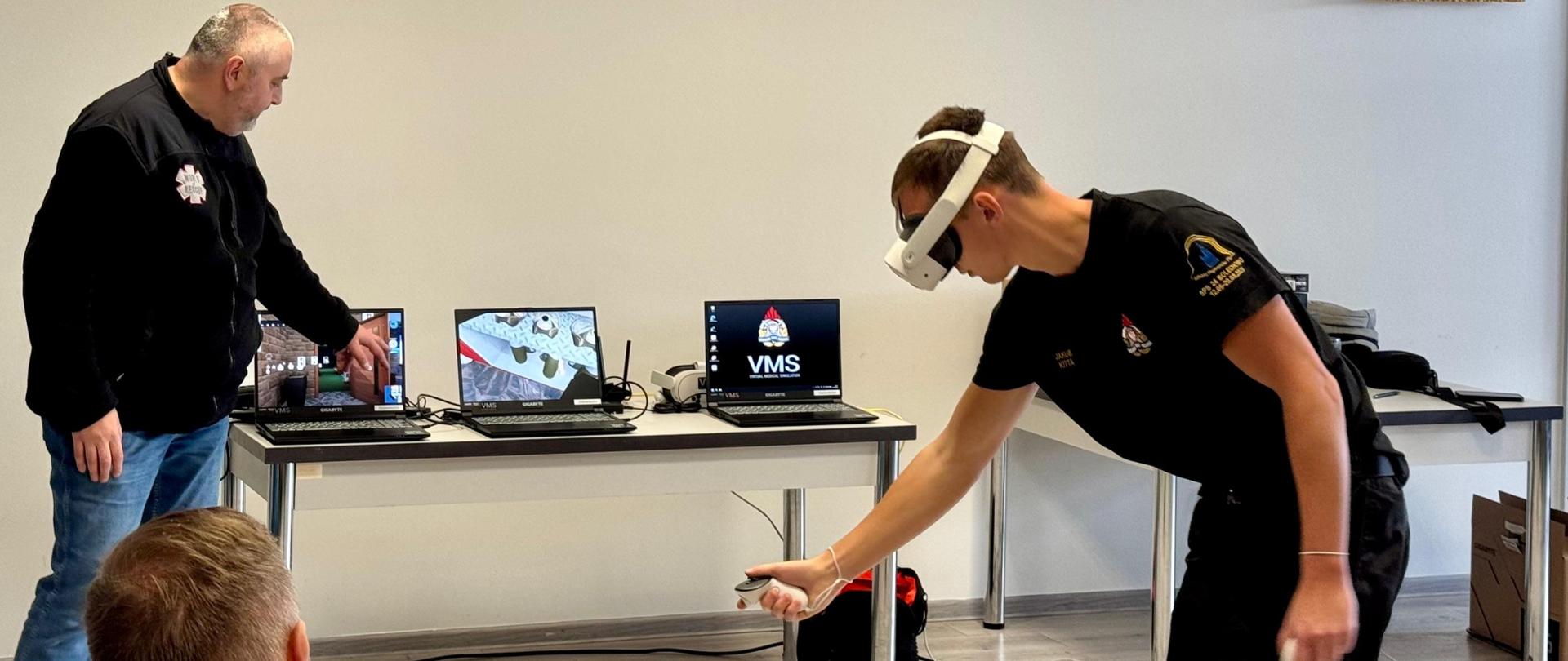 Zdjęcie zostało wykonane w remizie strażackiej. Zdjęcie przedstawia strażaka ćwiczącego w środowisku VR. W tle sprzęt oraz instruktor.