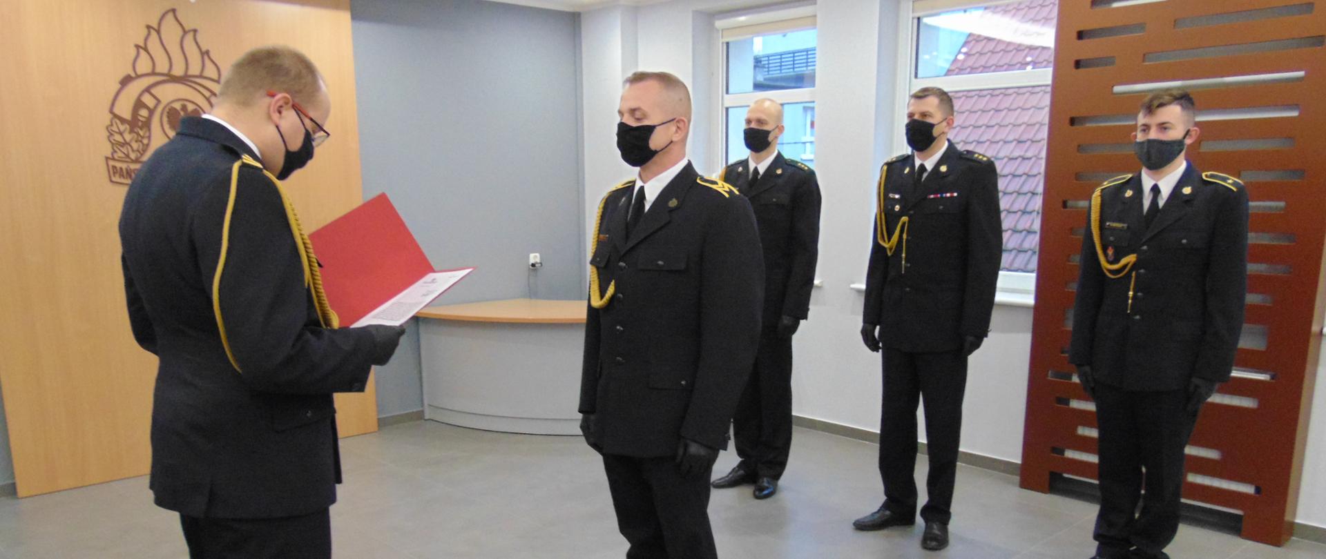 Uroczyste wręczenie nagrody przez zastępcę KM PSP Sopot . Strażacy stoja na swietlicty ubrani w mundury galowe Na środku sali. Komendant wręcza dokument nagrodzonemu. W tle stoi 4 strażaków oraz iwdoczne jest logo PSP na scianie.