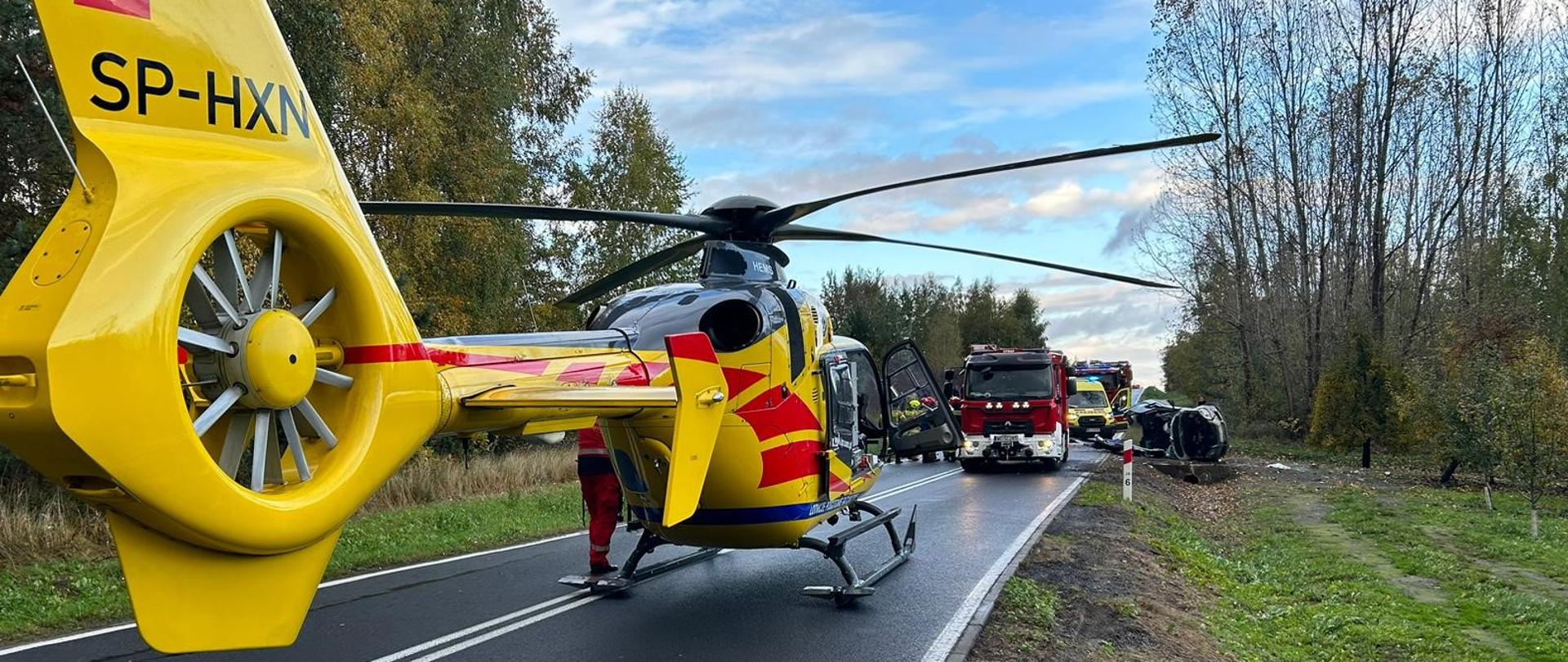 Zdjęcie przedstawia żółty śmigłowiec ratunkowy Lotniczego Pogotowia Ratunkowego (LPR) z numerem rejestracyjnym "SP-HXN", stojący na środku dwupasmowej asfaltowej drogi. Śmigłowiec ma czerwono-żółte pasy i jest w pełni gotowy do startu lub właśnie wylądował. W tle, w oddali, widać zgromadzone pojazdy ratunkowe: czerwony wóz strażacki z otwartymi drzwiami oraz karetkę pogotowia. Po prawej stronie drogi, tuż przy poboczu, leży przewrócony na bok, ciemny samochód osobowy, co wskazuje na miejsce wypadku drogowego. Obie strony drogi otoczone są drzewami i krzewami z jesiennym ulistnieniem. Niebo jest częściowo zachmurzone, ale z widocznymi fragmentami błękitu. Cała scena sugeruje trwającą akcję ratunkową po wypadku, z udziałem służb naziemnych i lotniczych.