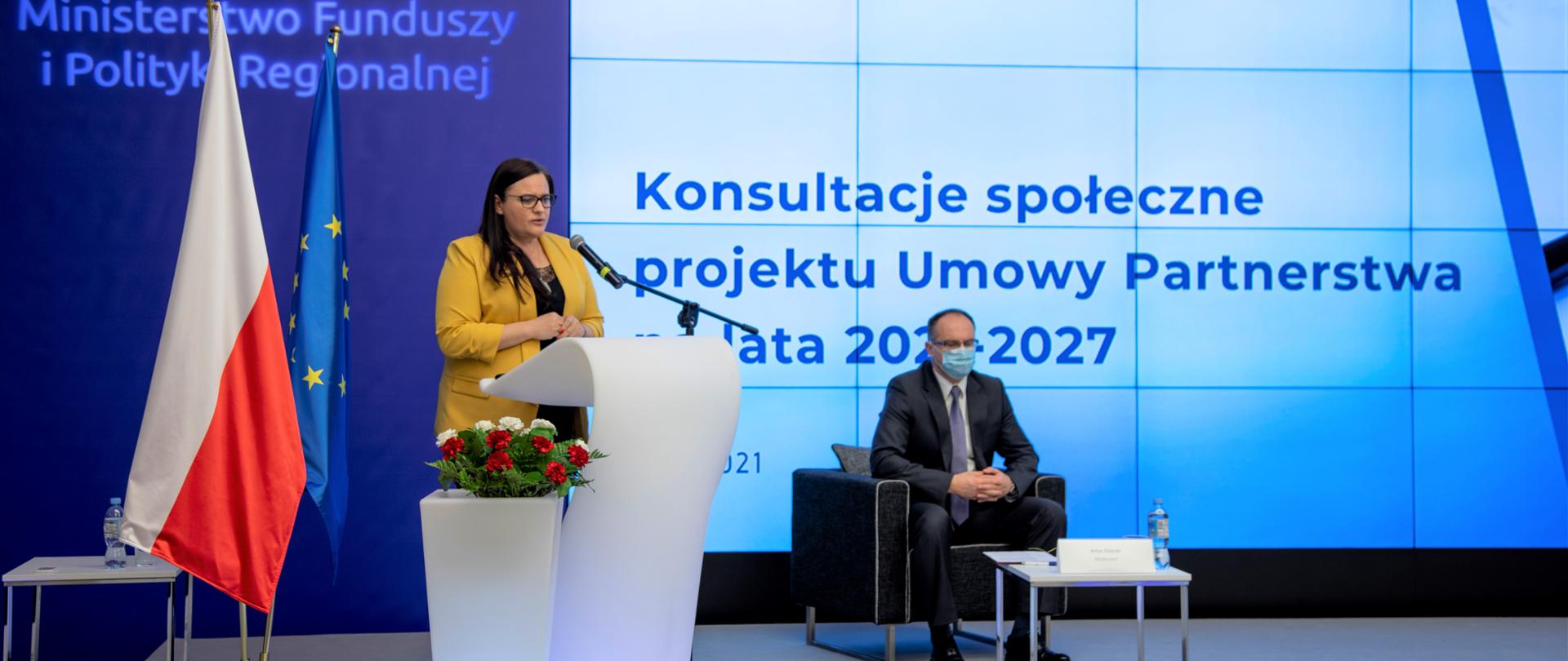 W mównicy z mikrofonem stoi wiceminister Małgorzata Jarosińska-Jedynak. Obok kwiaty w donicy. Z tyłu ścianka z napisem: "Ministerstwo Funduszy i Polityki Regionalnej" oraz dwie flagi PL i UE. Po prawej w fotelu przed stolikiem siedzi mężczyzna w maseczce. Za nim na ekranie napis: "Konsultacje społeczne projektu Umowy Partnerstwa na lata 2021-2027".
