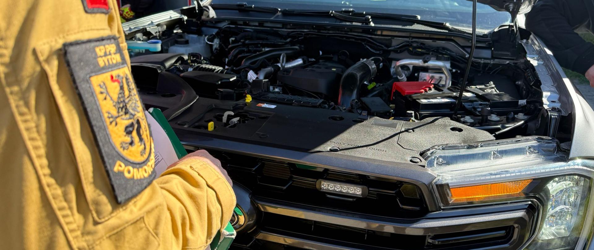 Zbliżenie na otwartą maskę srebrnego samochodu terenowego. Strażak w piaskowym mundurze z naszywką „KP PSP BYTÓW” stoi obok, trzymając podkładkę z dokumentami i dokonując inspekcji silnika. Widać detale osprzętu pod maską oraz fragment tablicy rejestracyjnej.