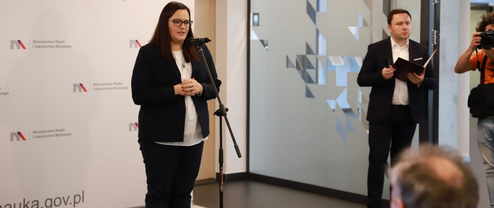 Podczas konferencji wiceminister Małgorzata Jarosińska-Jedynak mówi do stojącego przed nią mikrofonu. Za jej plecami ścianka z napisem Ministerstwo Nauki i Szkolnictwa Wyższego. Z prawej strony widać dwóch mężczyzn - fotografa oraz prowadzącego wydarzenie.