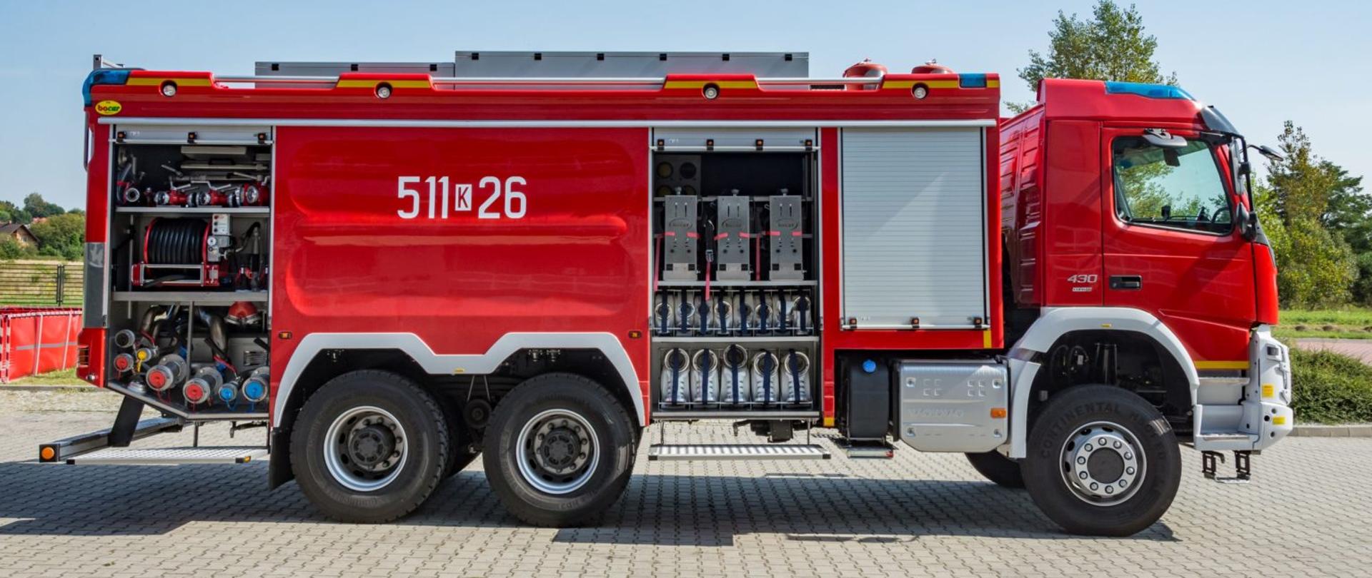Duży czerwony pojazd gaśniczy na tle niebieskiego nieba. Obok widoczny zbiornik wodny. Pojazd jest przedstawiony z boku, widać numer operacyjny pojazdu 511-26 . Samochód ma otwarte skrytki boczne, widoczny jest sprzęt pożarniczy, armatura wodno pianowa