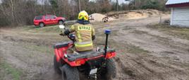Strażak w piaskowym ubraniu specjalnym na quadzie/ Quad czerwony z sygnalizacją ostrzegawczą. Pora dnia – dzień. W tle zabudowania i las oraz samochód SLRR, czerwony. Zachmurzenie duże.