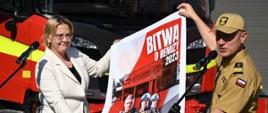 Z lewej Pani Minister Anna Moskwa w białym żakiecie i spodniach, z prawej Komendant Główny PSP w żółtym mundurze służbowym trzymający plakat akcji Bitwa o Remizy. W tle pojazdy pożarnicze ciężarowe koloru czerwonego
