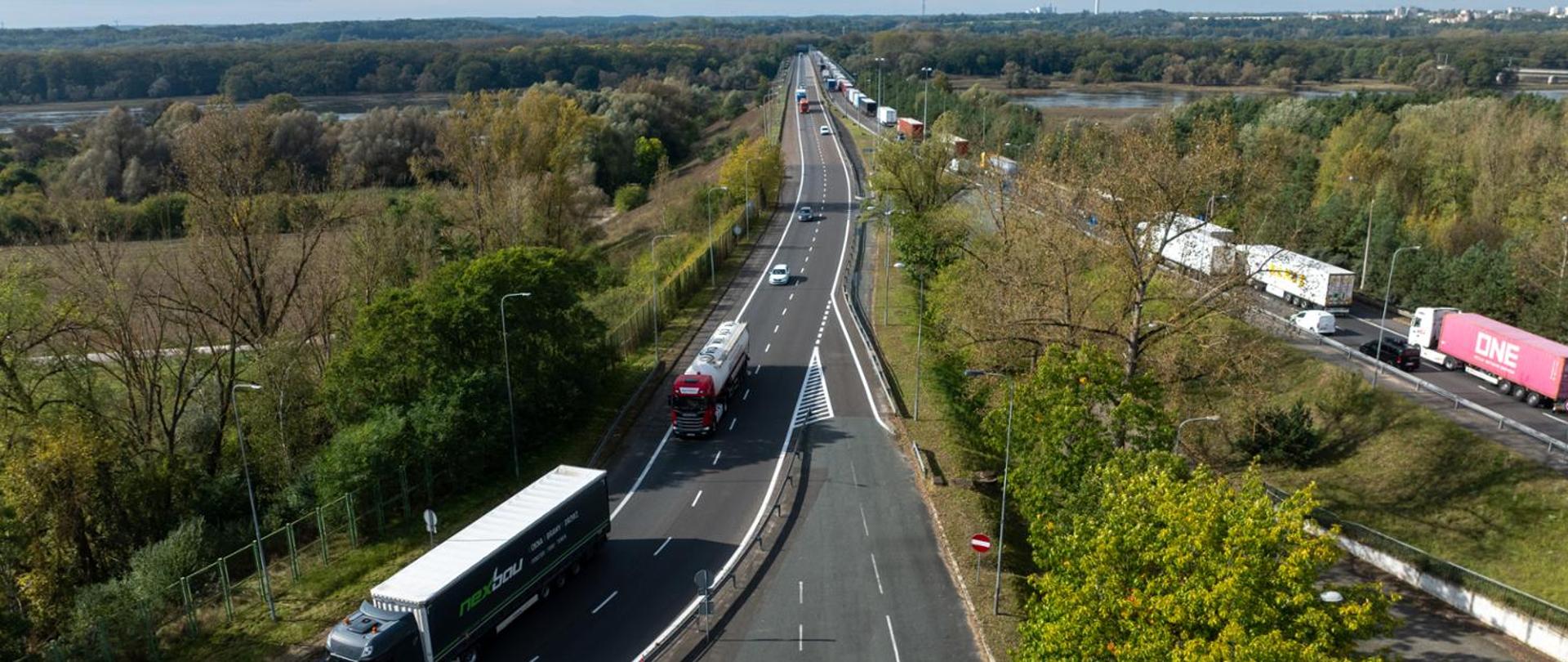 Widok z góry na autostradę z kilkoma ciężarówkami i samochodami, otoczoną zielenią i drzewami, w tle rozciąga się krajobraz z lasem i rzeką pod częściowo zachmurzonym niebem.