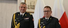 Pomorski komendant wojewódzki Państwowej Straży Pożarnej stoi obok strażaka trzymającego czerwoną teczkę. Za funkcjonariuszami znajdują się flagi państwowe. 