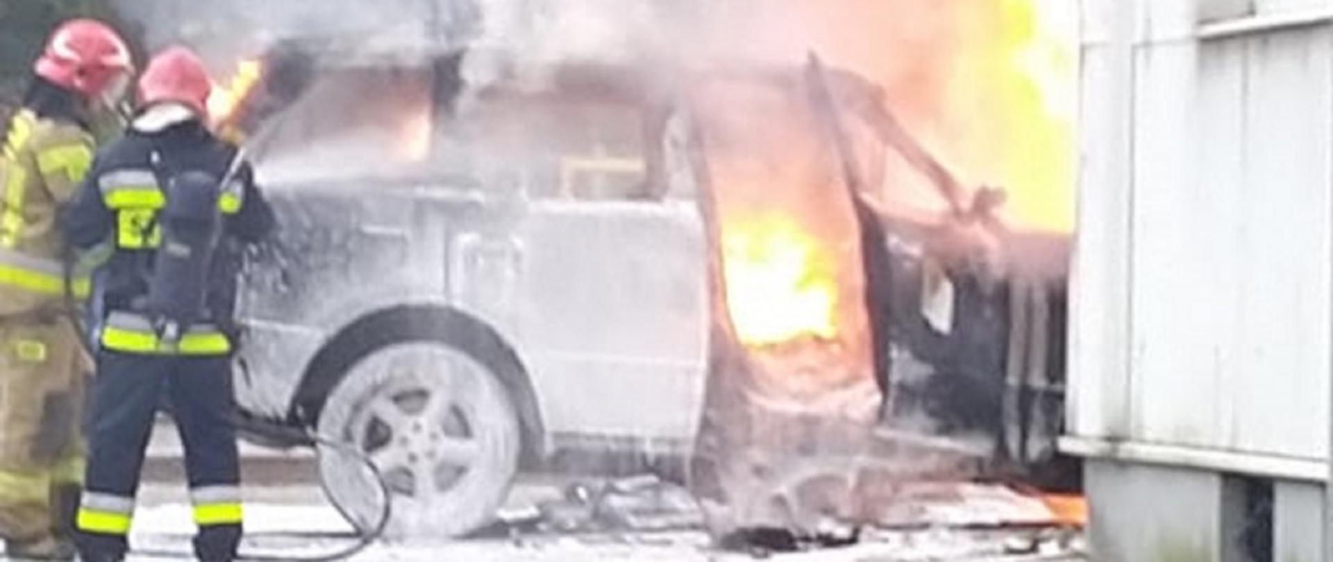 Na zdjęciu widoczny pożar samochodu osobowego oraz dwóch strażaków - jeden w jasnym, drugim w ciemnym ubraniu specjalnym. Jeden ma założony aparat powietrzny.