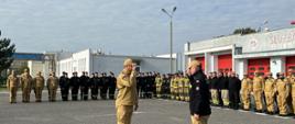 Strażak oddaje honor przed funkcjonariuszem Państwowej Straży Pożarnej ubranym w czarny polar obok stoją strażacy na baczność.