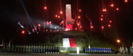 Na zdjęciu widać Pomnik Obrońców Wybrzeża w Gdańsku. Na niebie rozbłyskują czerwonym kolorem wojskowe flary. Na dole zdjęcia widać ustawione pododdziały wojska oraz harcerzy. Na środku fotografii widać biało czerwoną flagę.