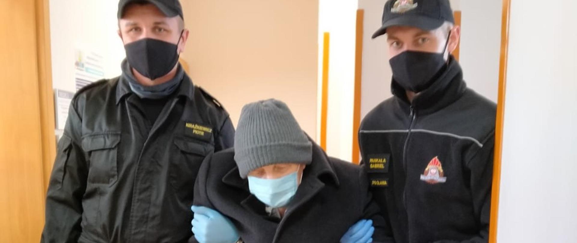 Na zdjęciu widać dwóch strażaków z oławskiej komendy. Pomiędzy nimi jest starszy mężczyzna, laską, podtrzymywany przez strażaków. Strażacy przywieźli seniora na szczepienie. 