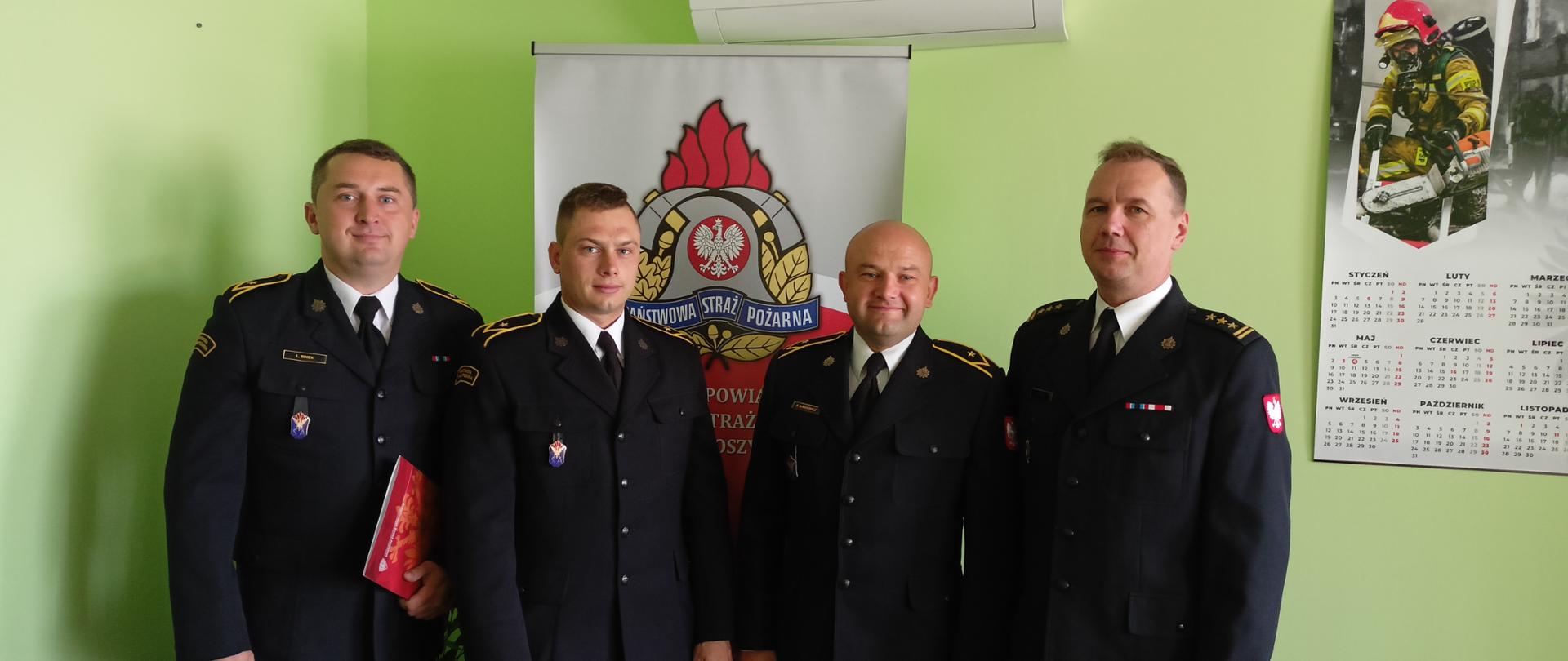 Zdjęcie przedstawia czterech strażaków w mundurach wyjściowych na tle baneru z logiem Państwowej Straży Pożarnej.