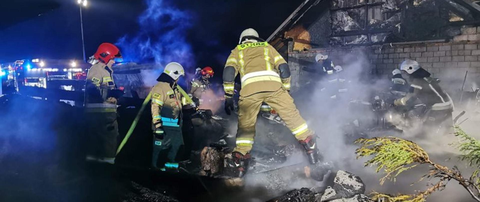 Jest pora nocna. Zdjęcie przedstawia spalony budynek gospodarczy. W tle widać nadpalony budynek mieszkalny. Strażacy pracują na terenie spalonego budynku gospodarczego przegrzebując pozostałości i przelewając wodą żarzące się elementy. Teren działań został oświetlony przez maszty oświetleniowe wozów bojowych które licznie przybyły na miejsce zdarzenia. Przez teren prowadzonej akcji poprowadzone są linie wężowe, które służyły do podania środka gaśniczego na palące się obiekty. 