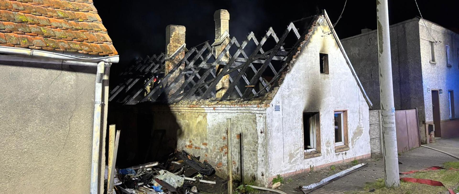 Na zdjęciu spalona więźba dachowa budynku mieszkalnego. Na poddaszu pracują strażacy wykonując prace rozbiórkowe. Teren oświetlają pojazdy pożarnicze. 
