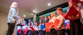 Na zdjęciu widać grupę dzieci i młodzieży, które występują na scenie podczas koncertu. Uczestnicy trzymają kolorowe śpiewniki, głównie w odcieniach czerwieni i niebieskiego. W centrum znajduje się dyrygentka, która prowadzi grupę. Ma na sobie jasną bluzkę w kolorowe wzory i stoi przodem do dzieci, które patrzą w jej stronę. Uczestnicy są ubrani w białe koszule oraz ciemne spódnice lub spodnie. Scena jest dobrze oświetlona, a tło ma zielony kolor, co tworzy przyjemną atmosferę. Całość wygląda na radosny i pełen energii występ.