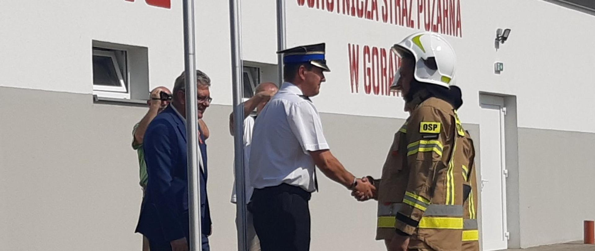 Strażak w białej koszuli gratuluje strażakom ubranym w umundurowanie bojowe. Za nim osoba cywilna ubrana w garnitur. W tle nowa remiza, na ścianie rysunek symbol słuchawki telefonicznej i napisy "112" oraz "Ochotnicza Straż Pożarna w Gorawinie.