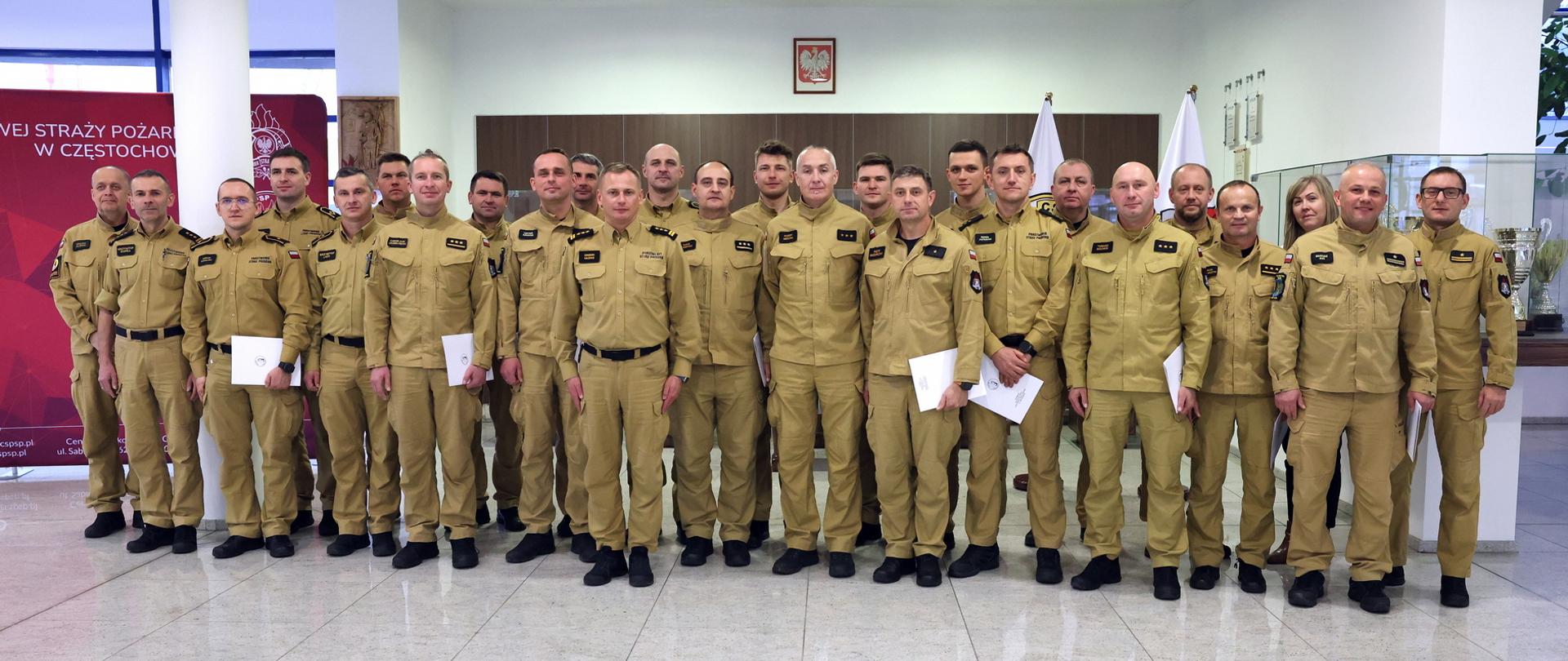 Pamiątkowe zdjęcie grupowe funkcjonariuszy PSP będących uczestnikami szkolenia z zakresu kierowania działaniami ratowniczymi na poziomie taktycznym (20 osób) w towarzystwie Zastępcy Komendanta Centralnej Szkoły PSP, kobiety oraz czterech oficerów CS PSP. Funkcjonariusze PSP w umundurowaniu służbowym koloru musztardowego