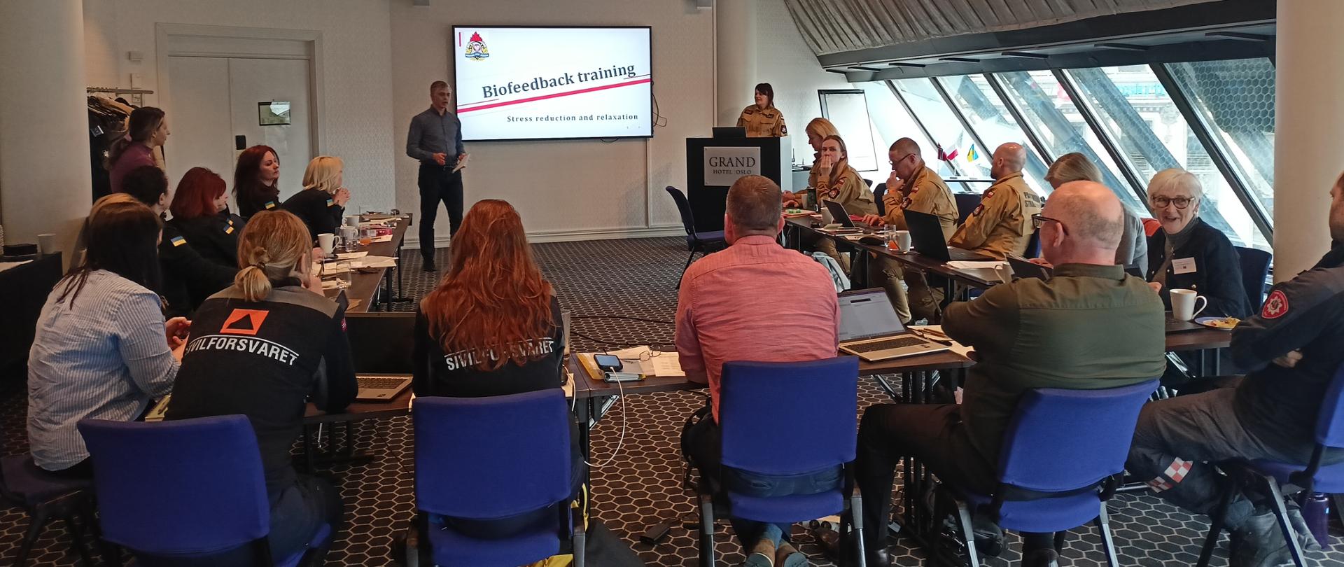 Kolejna część szkolenia, tym razem poświęcona treningowi biofeedback. Prelegent w cywilnym ubraniu omawia tematykę redukcji stresu i relaksacji. Na ekranie wyświetlony jest slajd z tytułem "Biofeedback training".