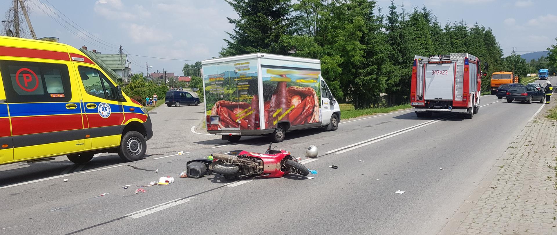Motocykl leży na środku drogi. Na drodze stoi karetka samochód dostawczy oraz samochód pożarniczy.