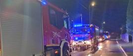 Zdjęcia w porze nocnej. Widać pojazdy straży pożarnej przed posesją gdzie doszło do pożaru. Sygnalizacja ostrzegawcza włączona. Droga publiczna. Lampy oświetlenia ulicznego włączone.
