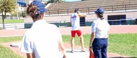 Mistrzostwa Europy Policjantów i Strażaków . Zdjęcie przedstawia fragment stadionu sportowego, na którym znajduje sie rzutnia do pchnięcia kulą. W kole stoi zawodnik przygotowujący sie do pchnięcia. Ma na sobie biała koszulkę z czerwonym napisem POLAND oraz czerwone spodenki. Za nim stoja dwie sędziny, jedna z nich trzyma czerwona chorągiewkę, druga - taśmę mierniczą. W tle trybuny.