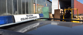 Dach samochodu Służby Celno-Skarbowej, w tle funkcjonariusz w wózku widłowym rozładowuje ciężarówkę z papierosami.