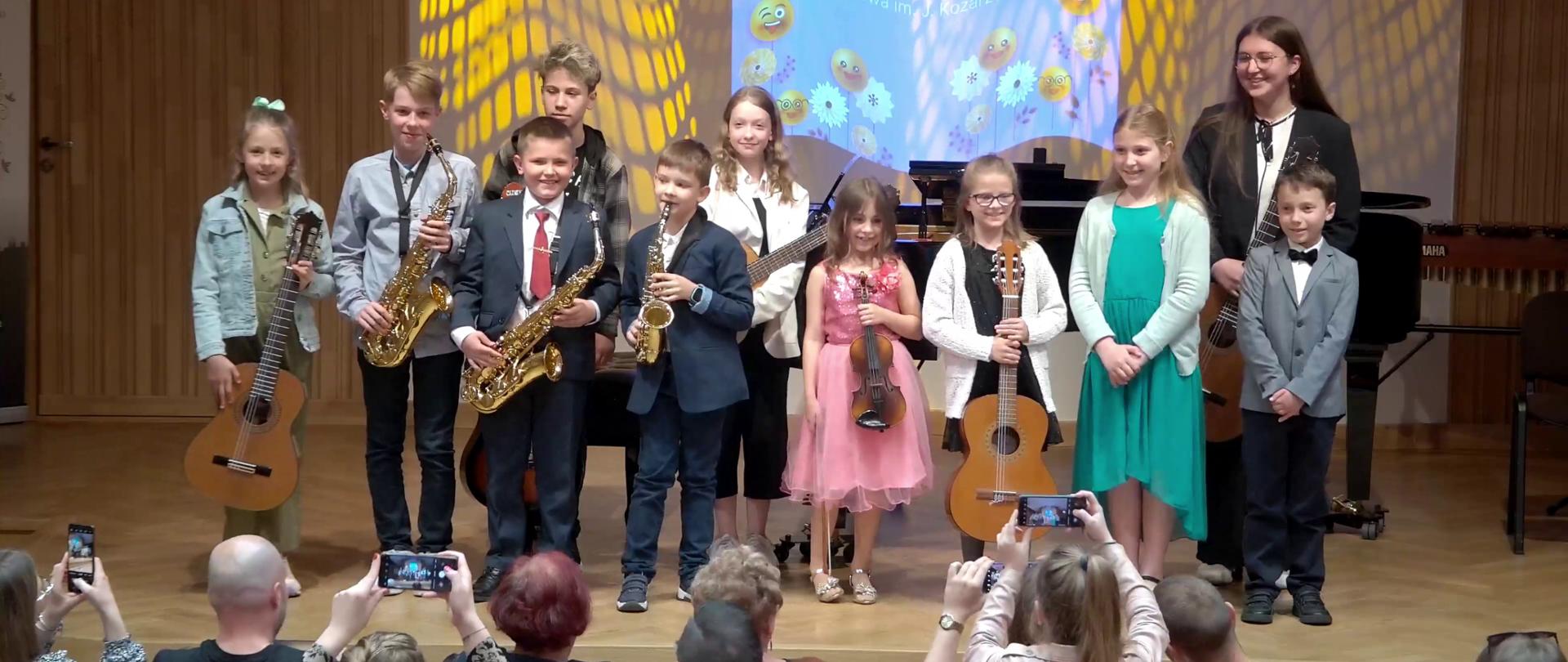 Zdjęcie. na estradzie stoją młodzi wykonawcy koncertu. Pozują do zdjęcia, które robią słuchacze koncertu. Część z nich trzyma swoje instrumenty. Za nimi stoi czarny fortepian. Uśmiechają się. W tle wyświetlony jest baner "Uśmiech dla taty i mamy".