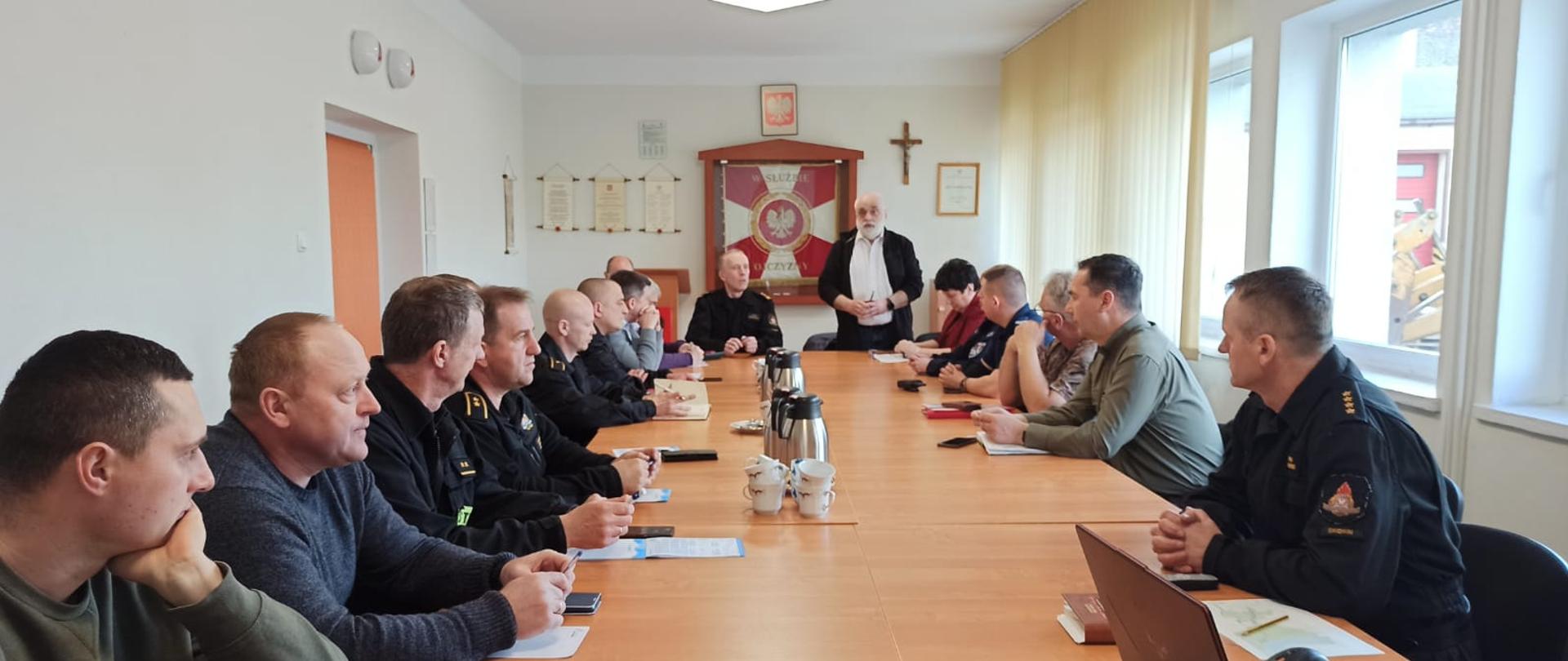 Zdjęcie przedstawia spotkanie, które odbywa się w sali szkoleniowej. W centrum zdjęcia widać stoły przy których siedzą funkcjonariusze oraz inni uczestniczący w szkoleniu. Jena osoba prowadząca szkolenie, która siedzi jest ubrana w niebieską koszulę. 