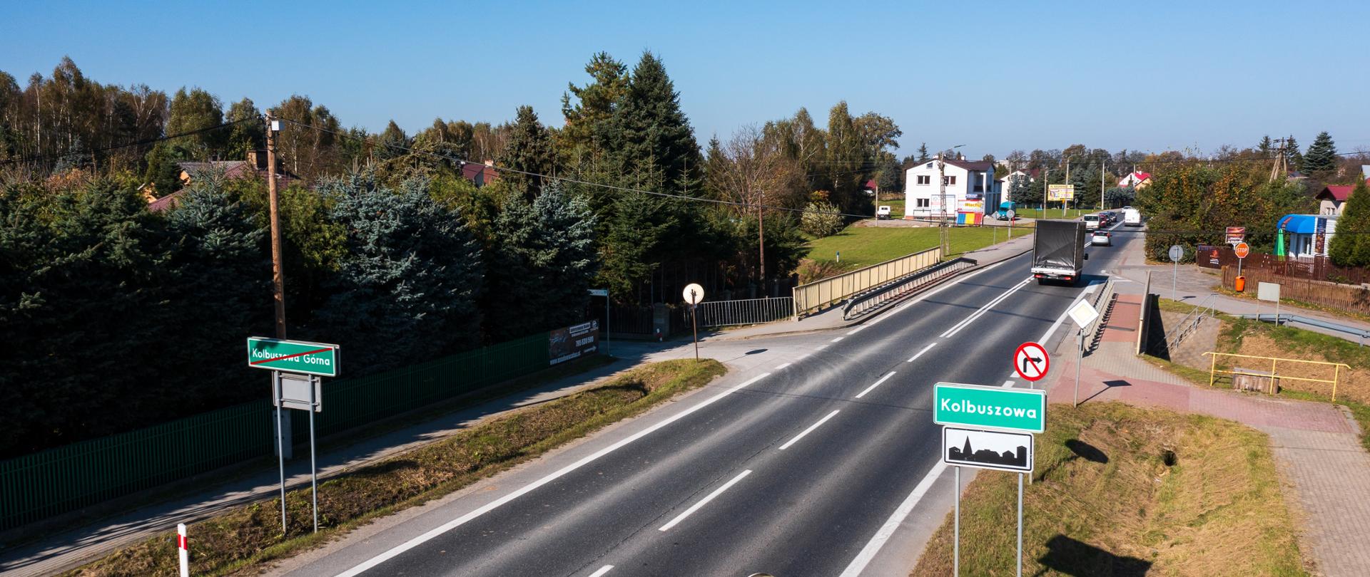 Na zdjęciu droga krajowa nr 9. Widać tablicę z nazwą miejscowości; widać chodnik, na drodze jedzie samochód, jest słonecznie