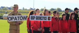 Uczestnicy zawodów strażackich stoją na murawie podczas zbiórki. Na pierwszym planie widoczne są tablice „OPATÓW” i „OSP GIERCZYCE”, obok grupa w czerwonych strojach sportowych.