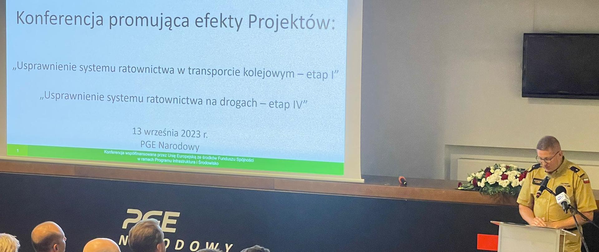 Zastępca komendanta głównego PSP przemawia na mównicy, na slajdzie napis Konferencja promująca efekty Projektów" usprawnienie systemu ratownictwa w transporcie kolejowym - etap I. Usprawnienie systemu ratownictwa na drogach - etap IV, 13 września 2023 PGE Narodowy