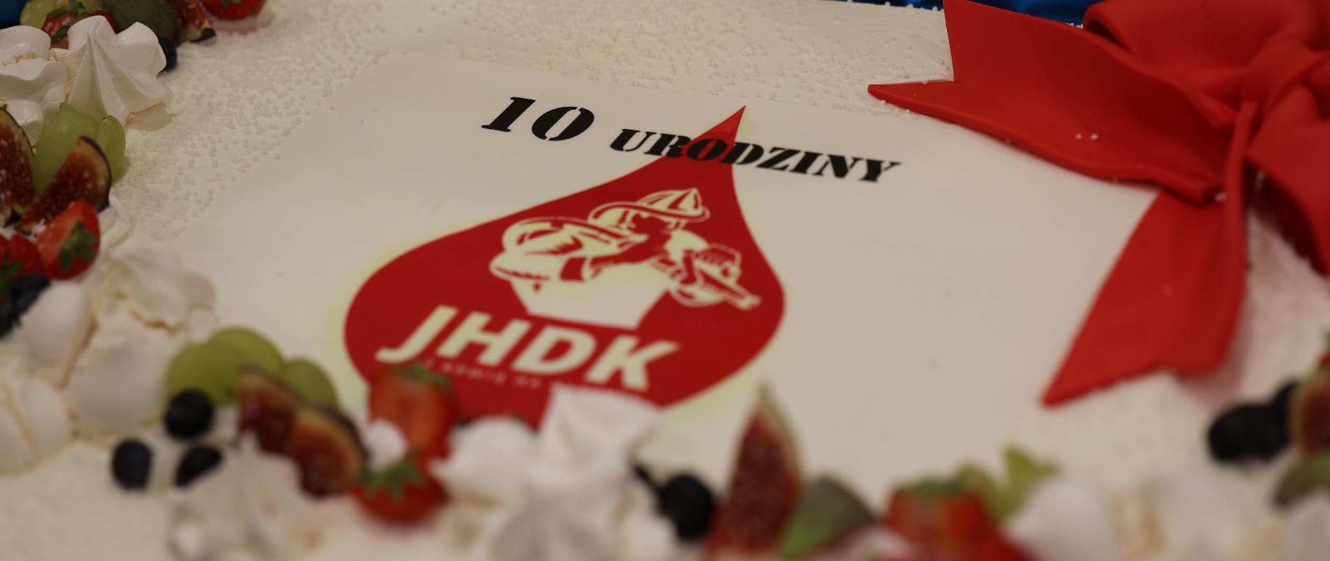 Jubileuszowy tort urodzinowy ozdobiony logo z napisem „10 URODZINY JHDK”, czerwoną wstążką oraz świeżymi borówkami i malinami