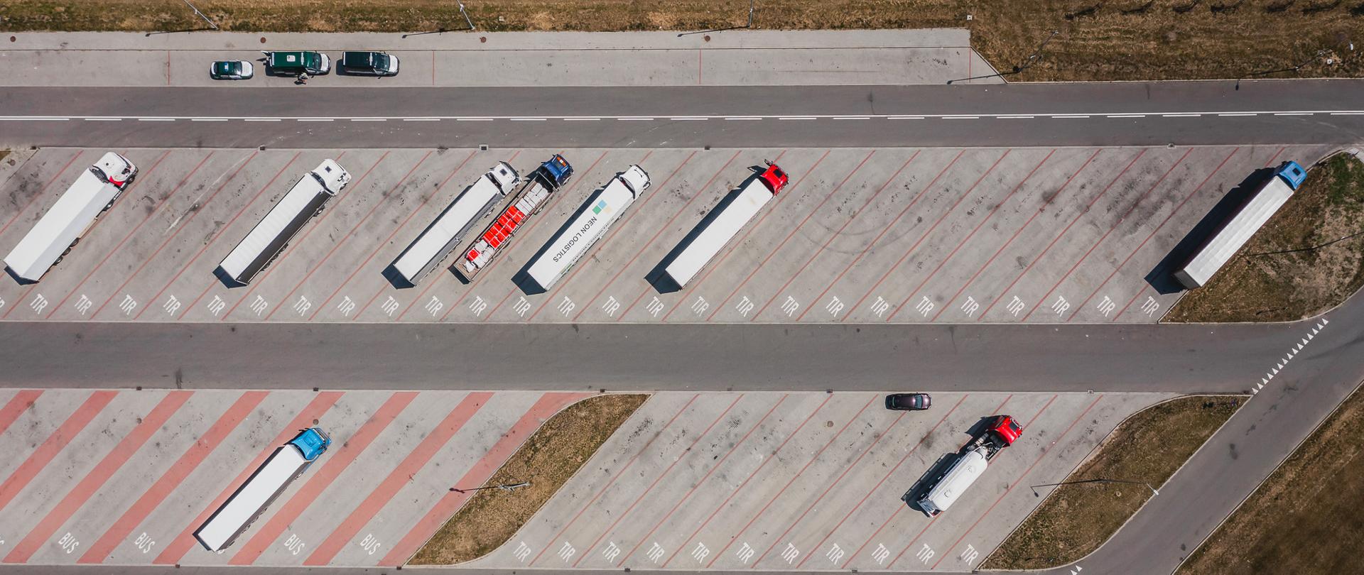 Widok z lotu ptaka na MOP Woźniki Zachód przy autostradzie A1 - zdjęcie poglądowe. Na wyznaczonych, skośnych stanowiskach oznaczonych napisem „TIR” stoją liczne białe i kolorowe zestawy ciężarowe. Pośrodku biegnie droga dojazdowa, a w dolnej części widoczne są miejsca parkingowe dla samochodów osobowych oraz wydzielone stanowiska oznaczone jako „BUS”. Całość otaczają pasy asfaltu, fragmenty suchej trawy i nieliczne nasadzenia drzew. Linie parkingowe są zaznaczone czerwonymi i białymi oznaczeniami.