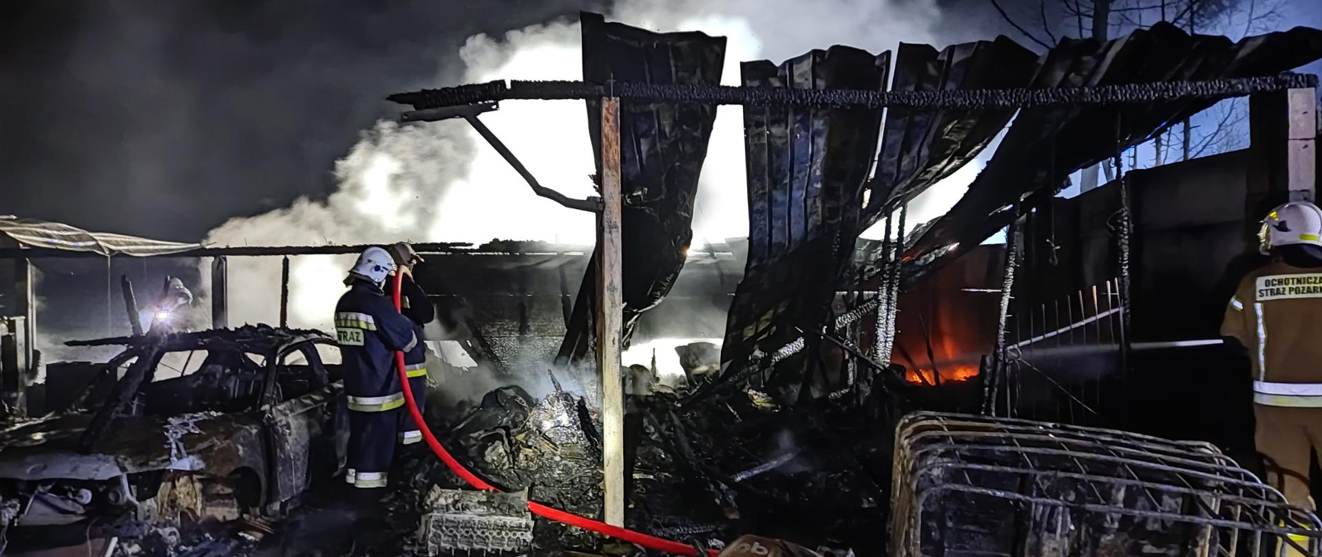 Zdjęcie przedstawia całkowicie spalony warsztat samochodowy. Na zdjęciu widać również spalony samochód oraz wyposażenie warsztatu. Strażak dogasza tlące się elementy wyposażenia warsztatu. Pora nocna. Nad warsztatem unosi się dym. 