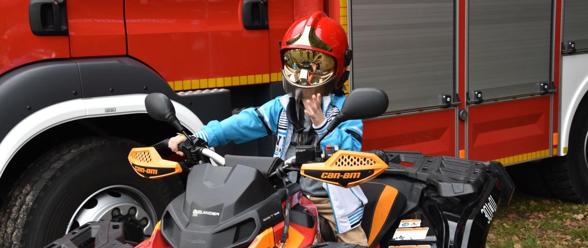 Na pierwszym palenie widzimy dziecko w hełmie strażackim na głowie siedzące na guadzie. Dziecko trzyma ręce na kierownicy. Quad stoi na tle samochodu pożarniczego.
