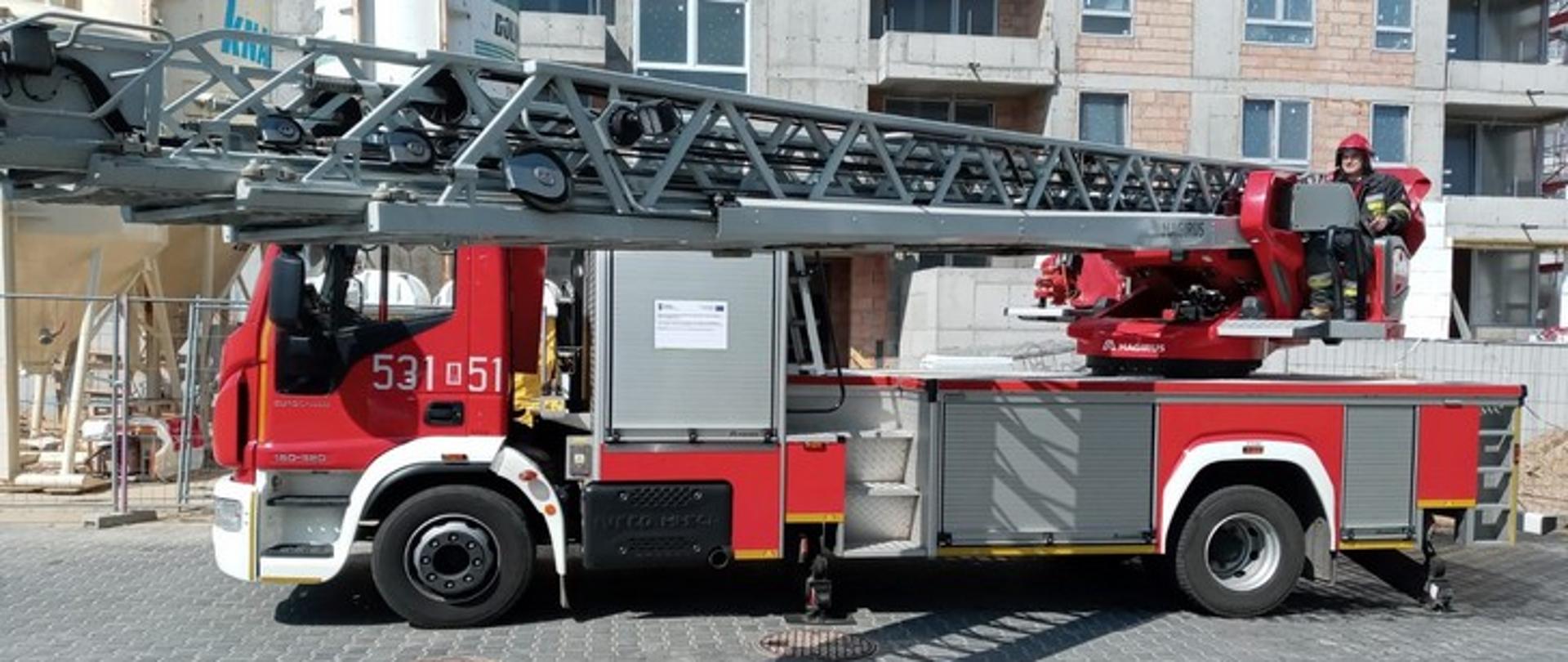 Obraz przedstawia strażacką drabinę mechaniczną z obsługującym ją strażakiem na tle nowo budowanego budynku wielorodzinnego.