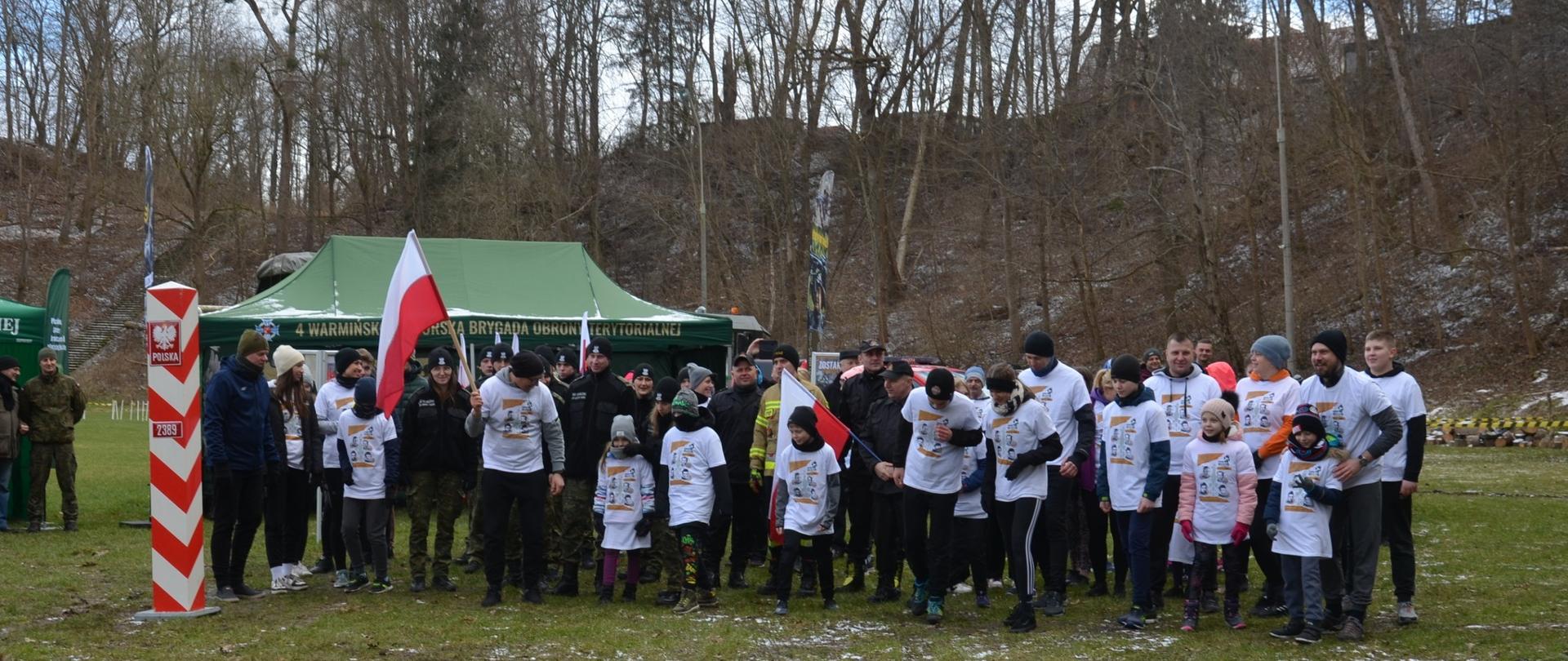 Na tle zielonej murawy boiska piłkarskiego. Stoi duża grupa ma starcie biegu, większość uczestników w białych koszulkach. Z lewej strony stoi symboliczny słup Graniczny.