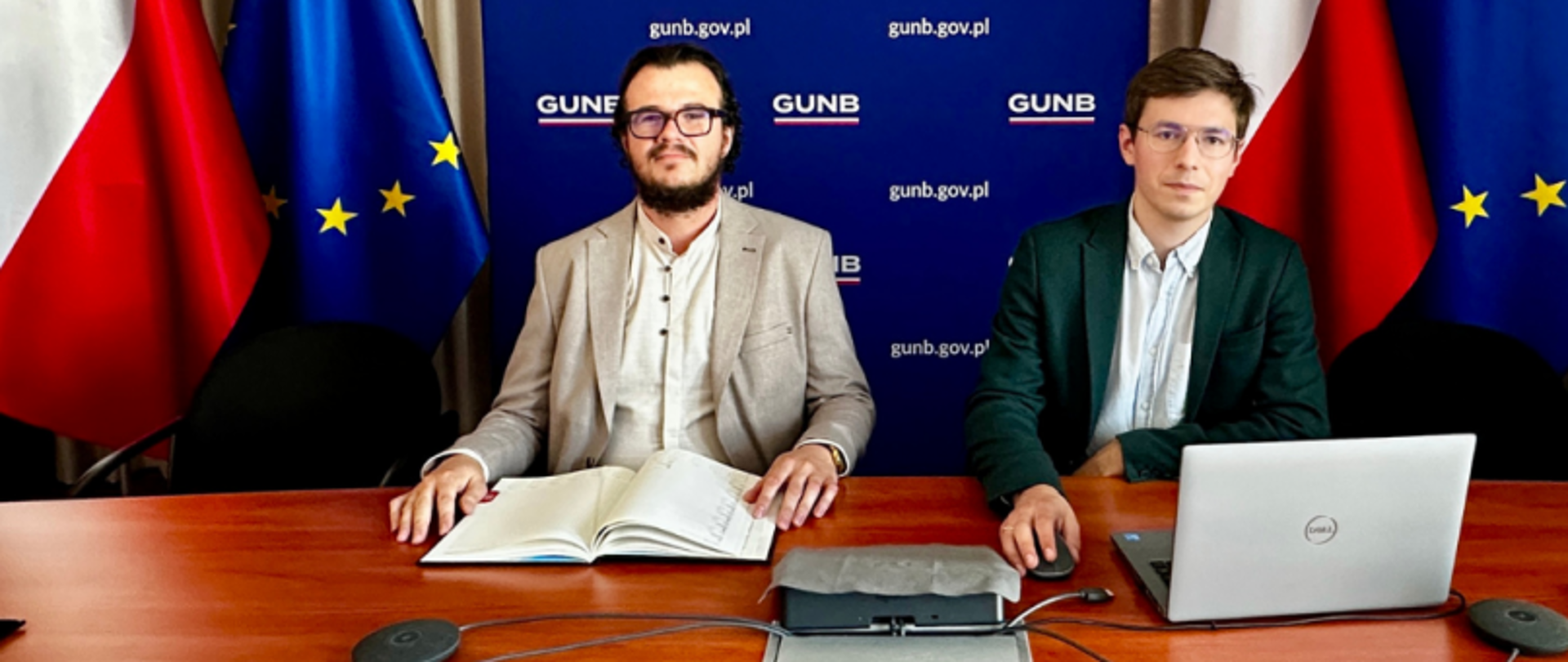 Filip Prusik-Serbinowski, Zastępca Głównego Inspektora Nadzoru Budowlanego (po prawej) oraz Piotr Centkowski, Zastępca Dyrektora Departamentu Orzecznictwa (po lewej), podczas prowadzenia szkolenia online. Siedzą przy stole z laptopem i dokumentami, w tle flagi Polski i Unii Europejskiej oraz ścianka z logotypem GUNB.