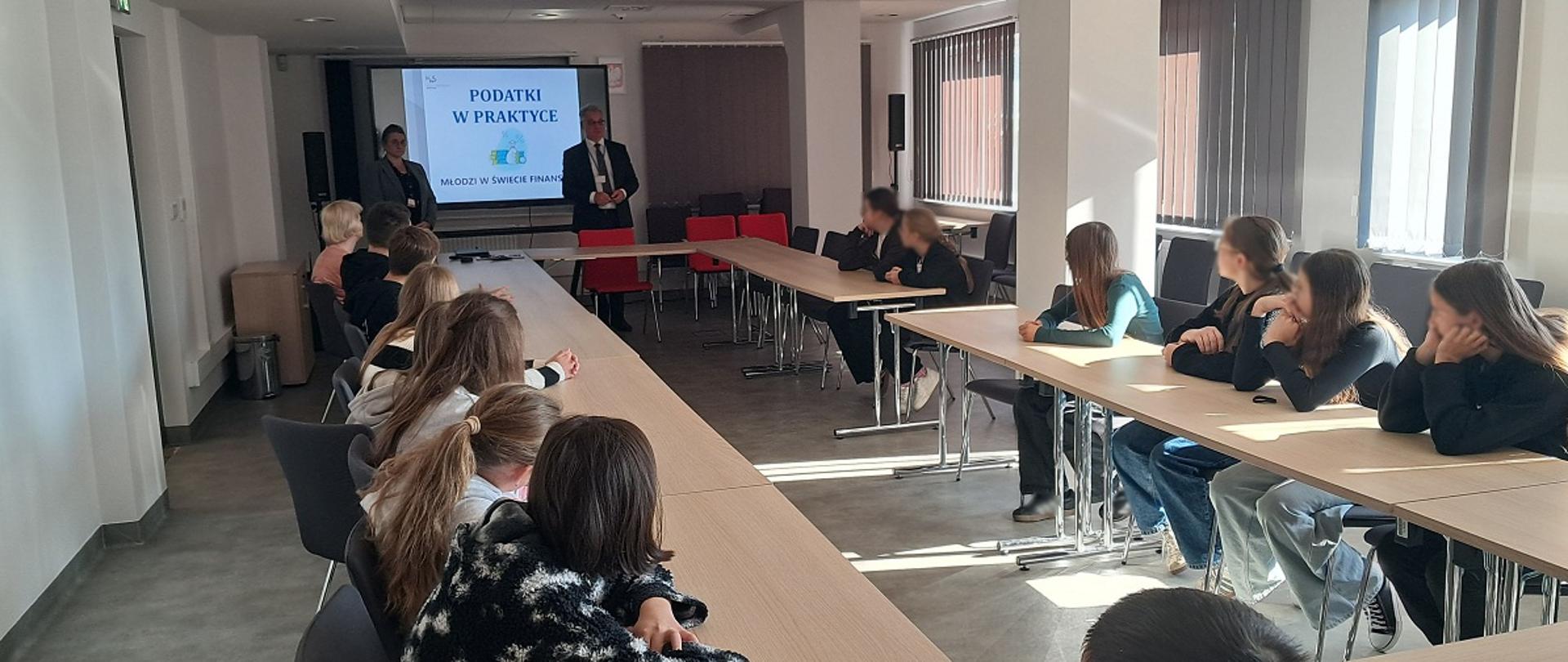 Sala konferencyjna. Uczniowie siedzą przy stołach. Naczelnik US w Sanoku wita młodzież. W tle wyświetla się prezentacja.
