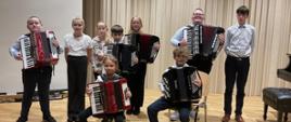 Grupa dzieci i nastolatków pozuje na scenie sali koncertowej z akordeonami. Część uczniów trzyma akordeony w rękach, a dwoje siedzi na krzesłach z instrumentami na kolanach. W tle znajdują się jasne, pionowe panele akustyczne oraz fortepian po prawej stronie. Wszyscy są ubrani odświętnie i patrzą w stronę aparatu.