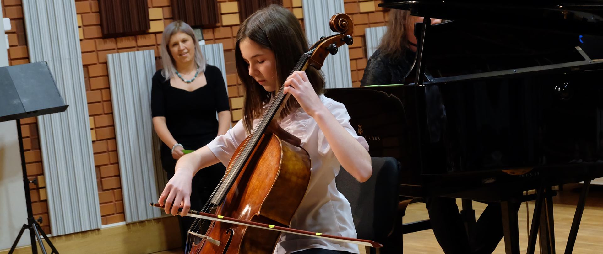 Dziewczynka gra na wiolonczeli, w tle siedzi jej mama i się uśmiecha.